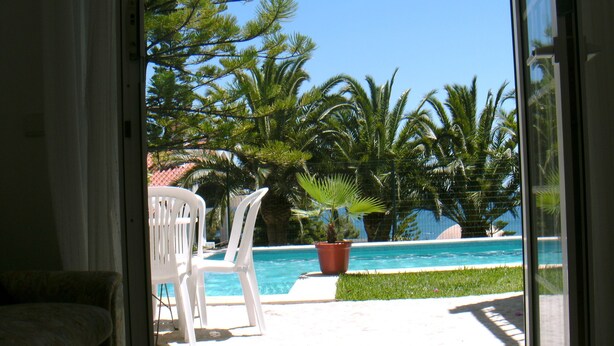 A view through a glass door to a pool area with chairs and a potted plant. - Apartment m. Pool u. Sea view, about 500m to the beach in Sesimbra for max. 4 pers (Sesimbra)