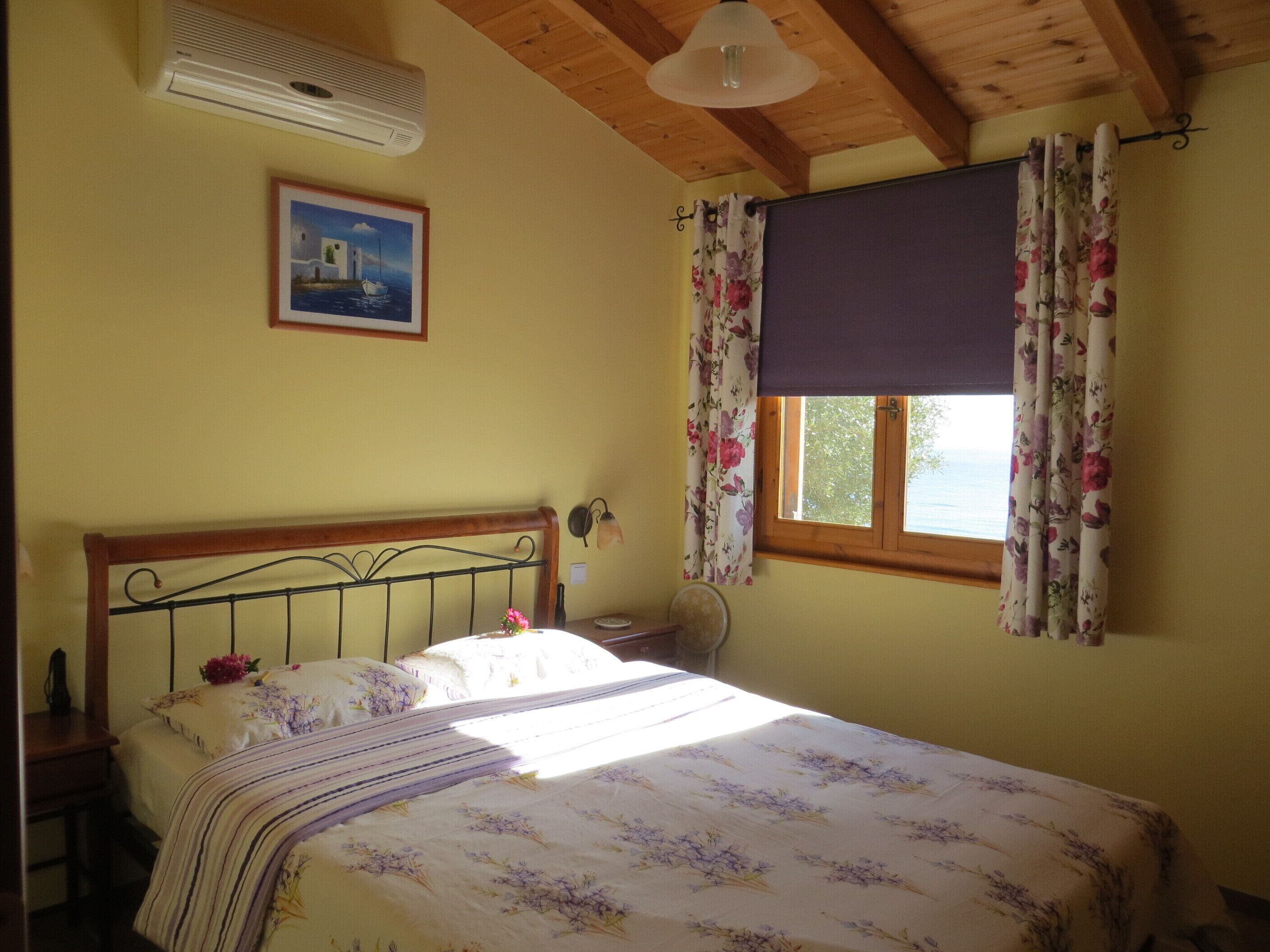 A bedroom with a bed, a window with curtains, a wall-mounted air conditioner, and a framed picture.