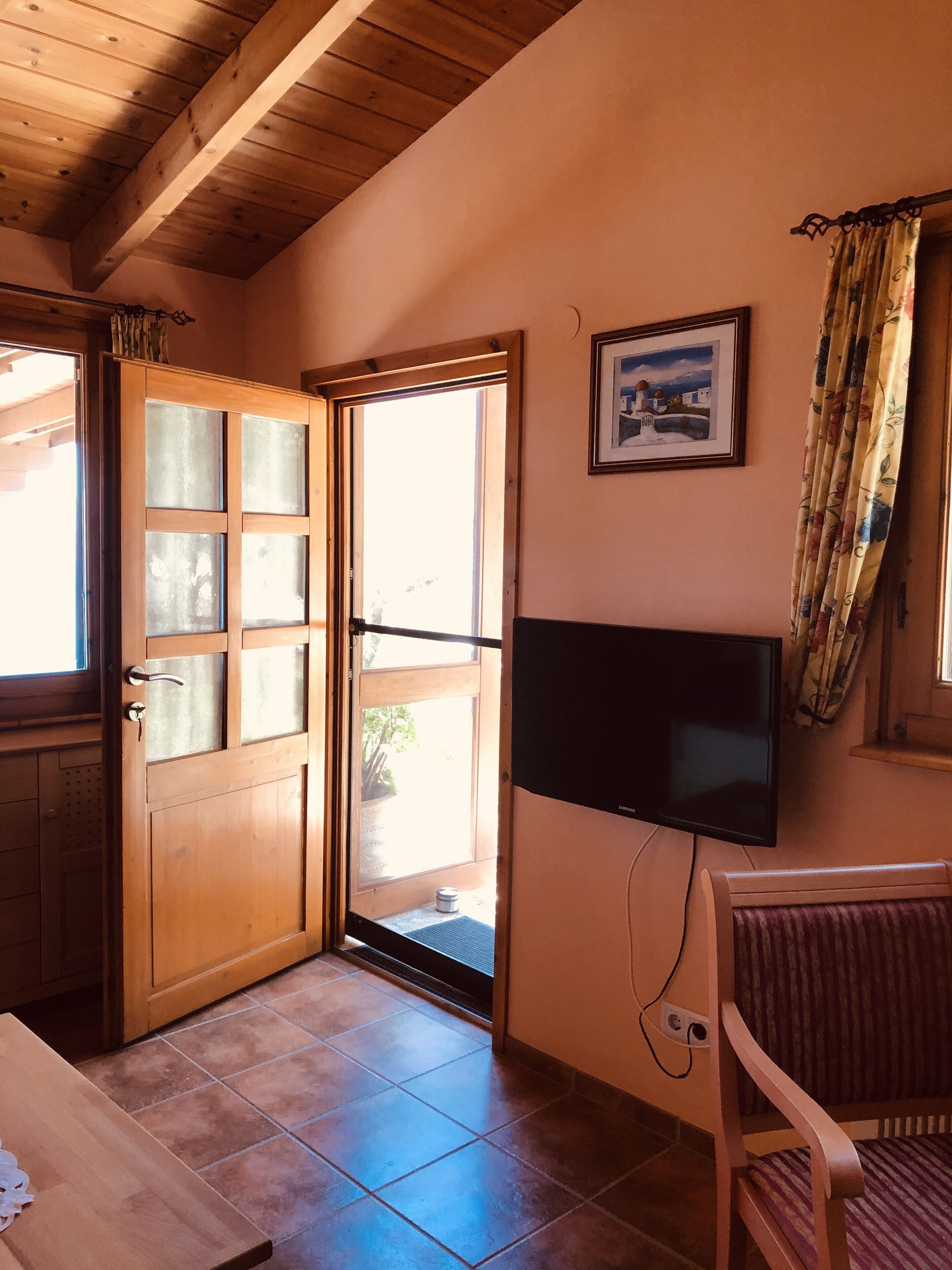 Una habitación con puerta de madera, televisor en la pared, una silla y ventana con cortinas.