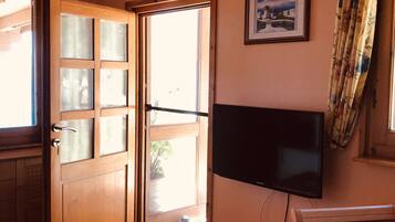 Una habitación con puerta de madera, televisor en la pared, una silla y ventana con cortinas.