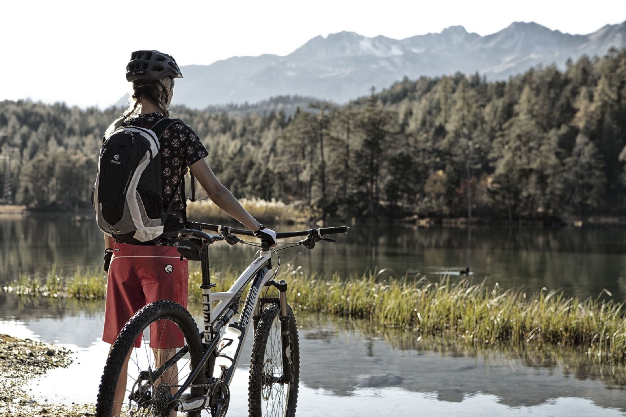 Ein Mountainbiker steht neben einem Fahrrad an einem See, mit Bergen im Hintergrund.