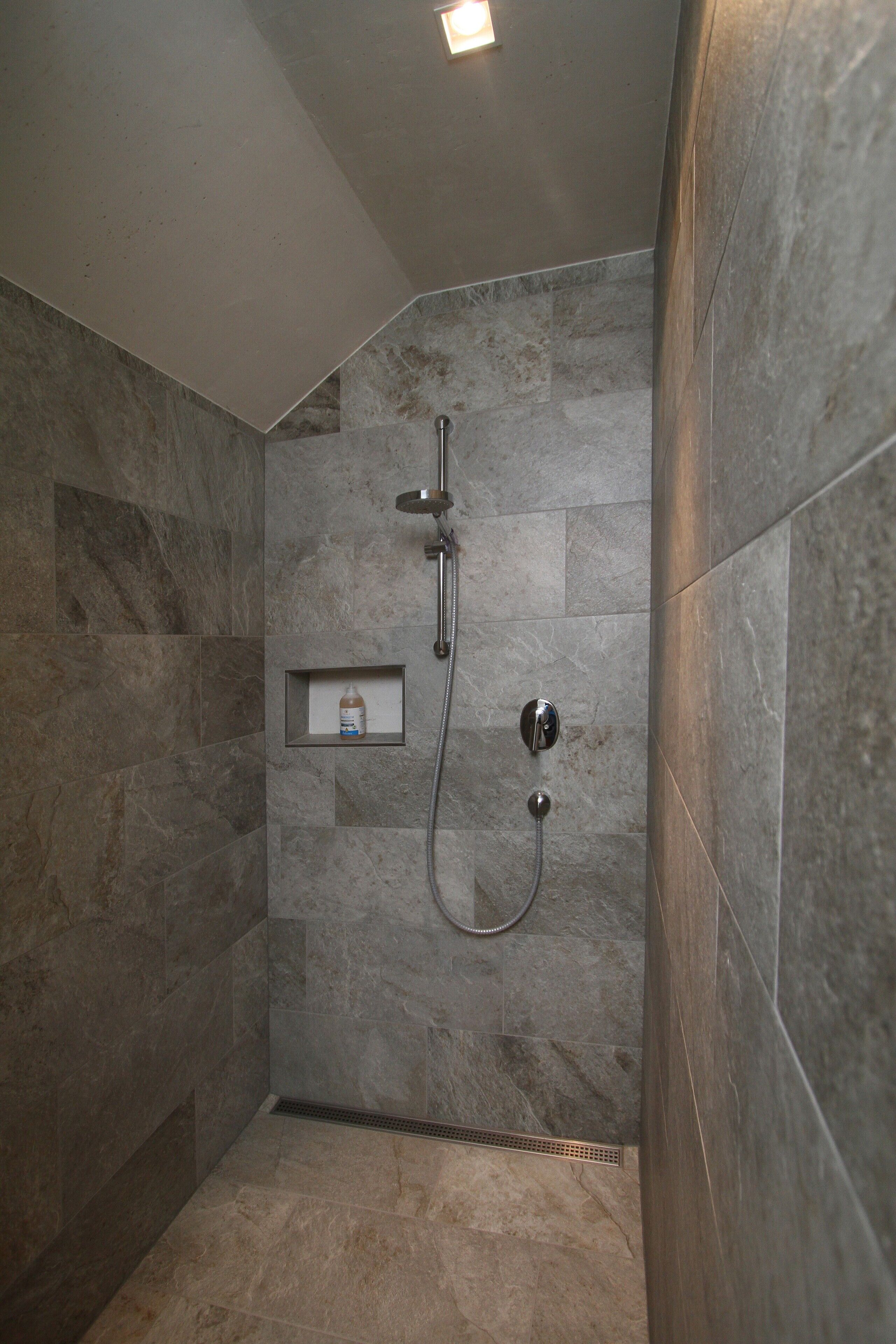 Une douche avec un pommeau de douche à main et un robinet mural, une petite étagère avec une bouteille, et une grille d’aération au sol.