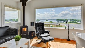 A cozy living room with a black leather chair, a white sofa, a small table with a plant, and a large window offering a view of a lake and trees.