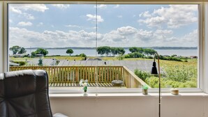 A room with a large window overlooking a lake, a wooden deck, and a chair.