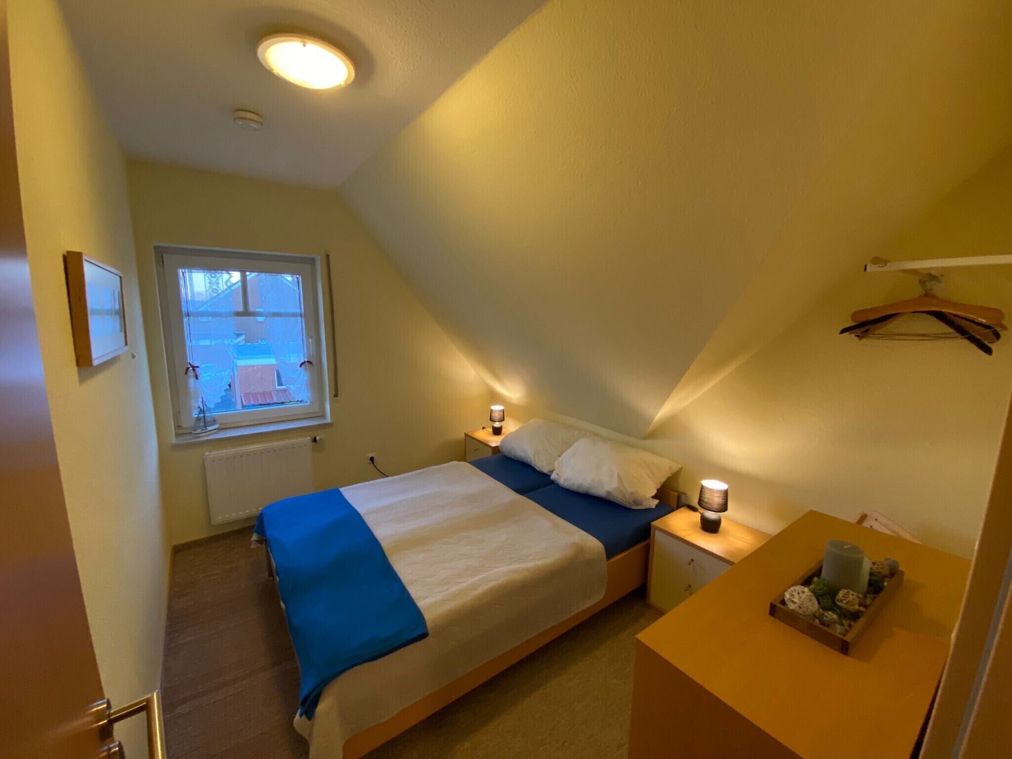 A small, well-lit bedroom with a single bed, a bedside table, a hanging coat rack, and a window with a view of the outside.