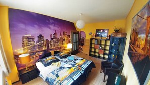 Een slaapkamer met een stadsmuurschildering, een bed met een beddegoed met stadsprint, een boekenplank en een bureau met een lamp.