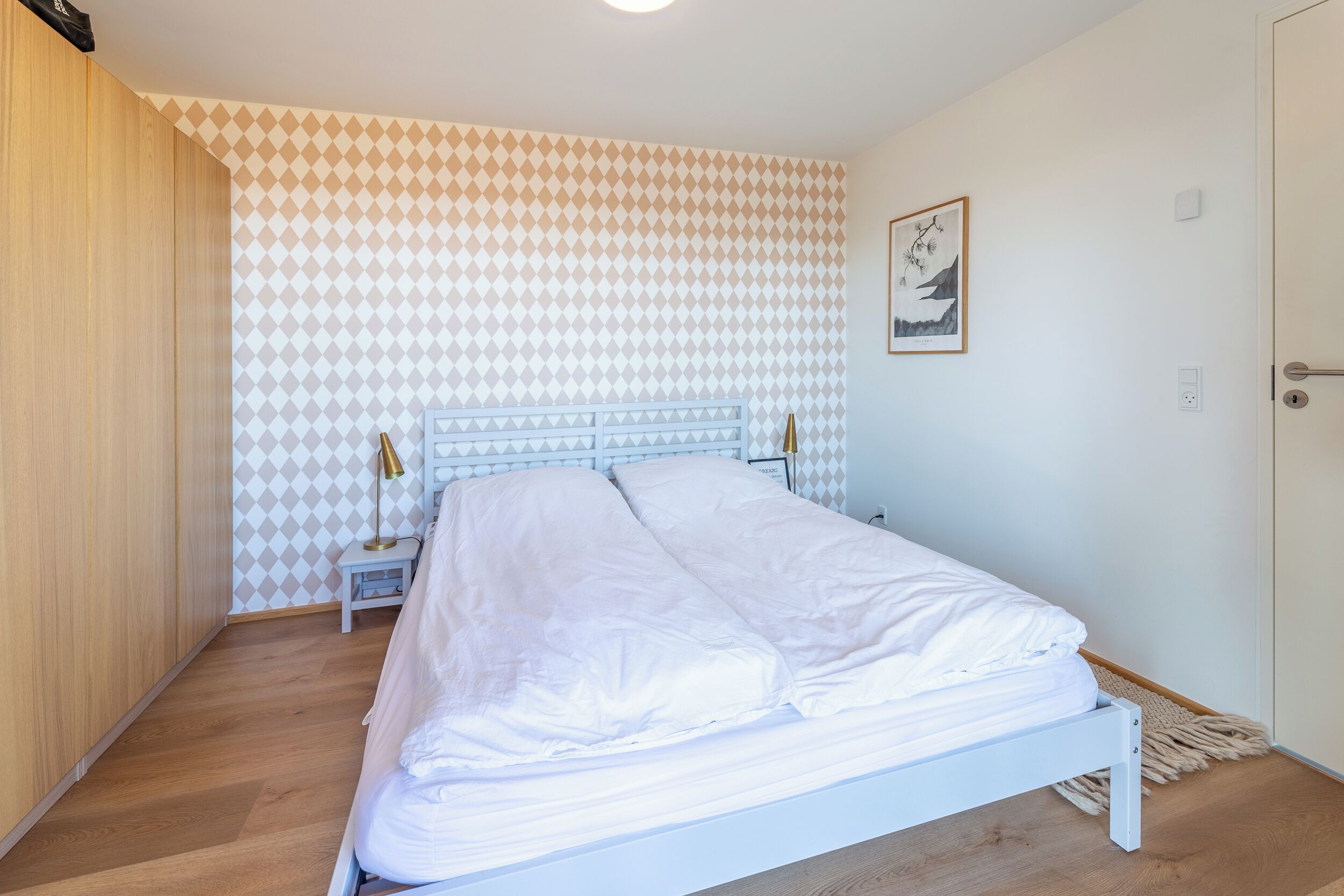 A bedroom with a bed, a nightstand, a lamp, and a wall with a geometric pattern.