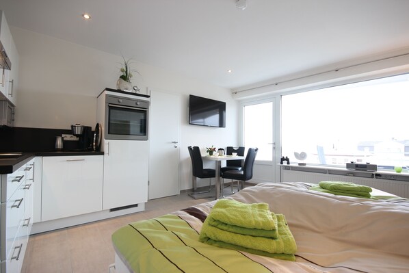A modern hotel room with a bed, a small kitchenette, a dining area, and a large window.