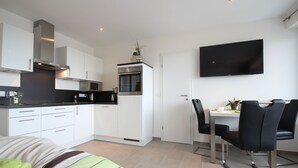 Une cuisine moderne avec des armoires blanches, une hotte de cuisinière en acier inoxydable et un four à micro-ondes. On y trouve également un coin repas avec une table et des chaises, ainsi qu’un téléviseur à écran plat fixé au mur.