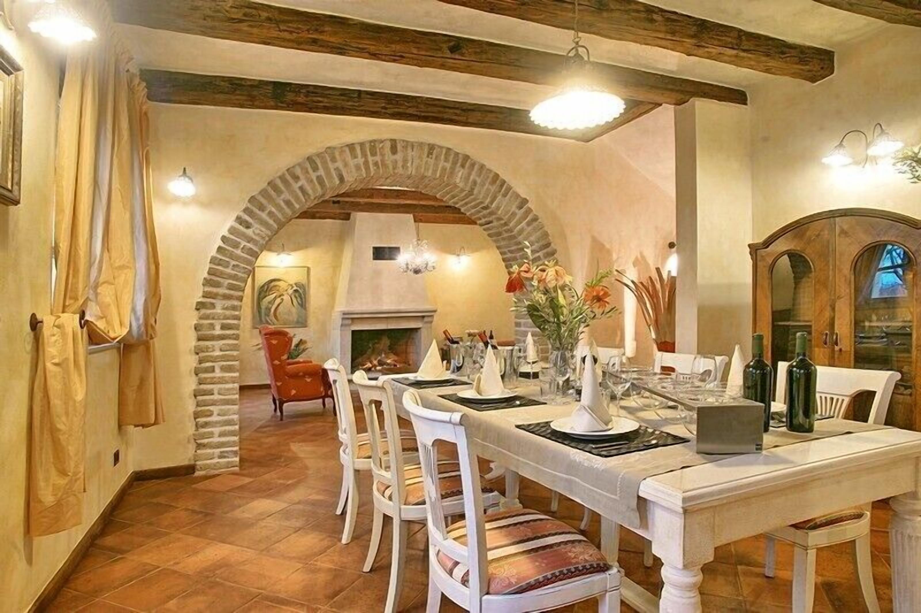 Ein Essbereich mit einem Steingewölbe, weißen Stühlen, einem langen Tisch, der für eine Mahlzeit gedeckt ist, und einem Kamin im Hintergrund.