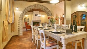 Un comedor con un arco de piedra, sillas blancas, una mesa larga dispuesta para una comida y una chimenea al fondo.