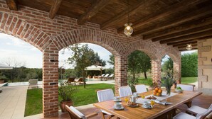 Un patio cubierto con un arco de ladrillo, una mesa de madera, sillas blancas y vistas a una piscina y jardín.