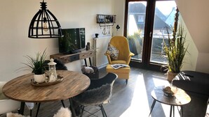 Un saló modern amb una taula de dinar de fusta, un sofà armchair groc i un canapè. Hi ha un televisor muntat a la paret, un radiador i una finestra amb vista a edificis.
