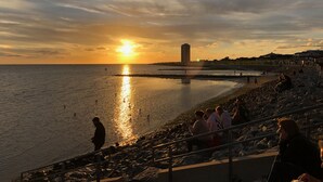 Vista d'un tramont a la costa, amb cel clar, un edifici llunyà i gent a la platja.
