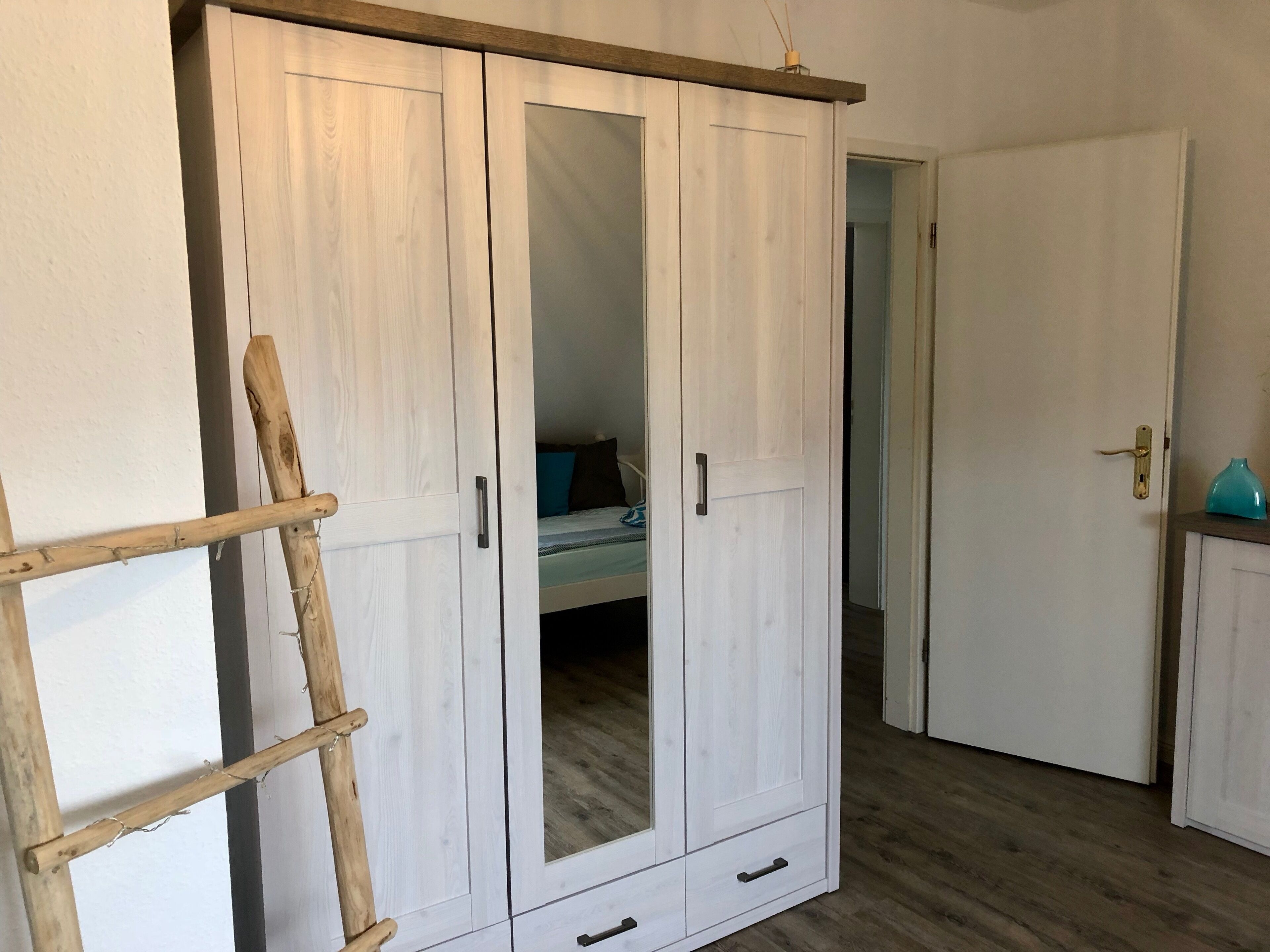 Ein Zimmer mit einem großen Kleiderschrank, einem Bett mit blauem Kissen und einer kleinen Kommode mit einer blauen Vase.