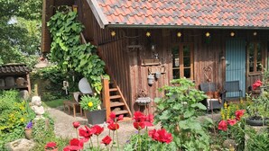 Una cabaña de madera rústica con techo de tejas, un porche de madera y un jardín con flores rojas.