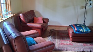 Una sala de estar con un sofá de cuero marrón, un reposapiés a juego y una alfombra roja. Se ve una ventana con cortinas, una lámpara de pared y un cuadro.