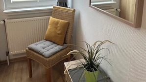 A room with a wicker chair, a small table with a plant, a mirror, and a window with blinds. - Apartment 3 with balcony, animal free  near the south beach (Borkum)