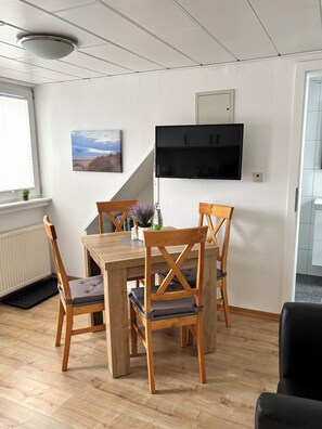 A dining area with a wooden table and chairs, a mounted TV, a window with blinds, and a radiator.