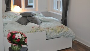 Un dormitorio con cama, ventana con cortinas, mesita de noche con un florero y una lámpara colgante.