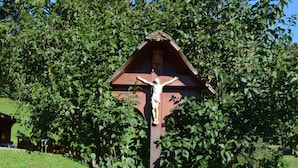 A cross with a figure, surrounded by greenery and a building in the background. - Freiburg, Southern Black Forest, Schauinsland Hofsgrund, farm houses, (Oberried)