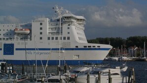 Un barco grande blanco y azul amarrado en un puerto deportivo, con varios botes más pequeños y un cielo despejado.
