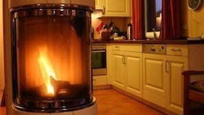 Una cocina moderna con una estufa de leña central, armarios blancos y un ventanal con cortinas rojas.