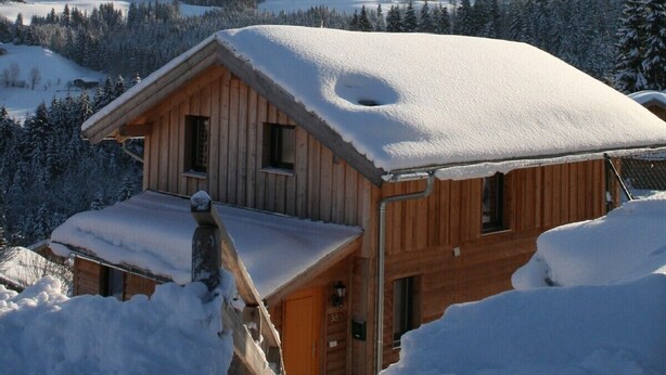 Una cabaña de madera con techo cubierto de nieve, rodeada por un paisaje nevado y montañas al fondo.