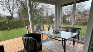 Une véranda dotée d’une porte vitrée, d’une terrasse en bois, d’une table avec des chaises et d’un banc.