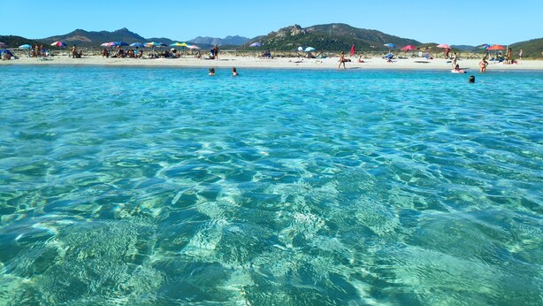 A beach with clear turquoise water, people swimming and sunbathing, and mountains in the background.