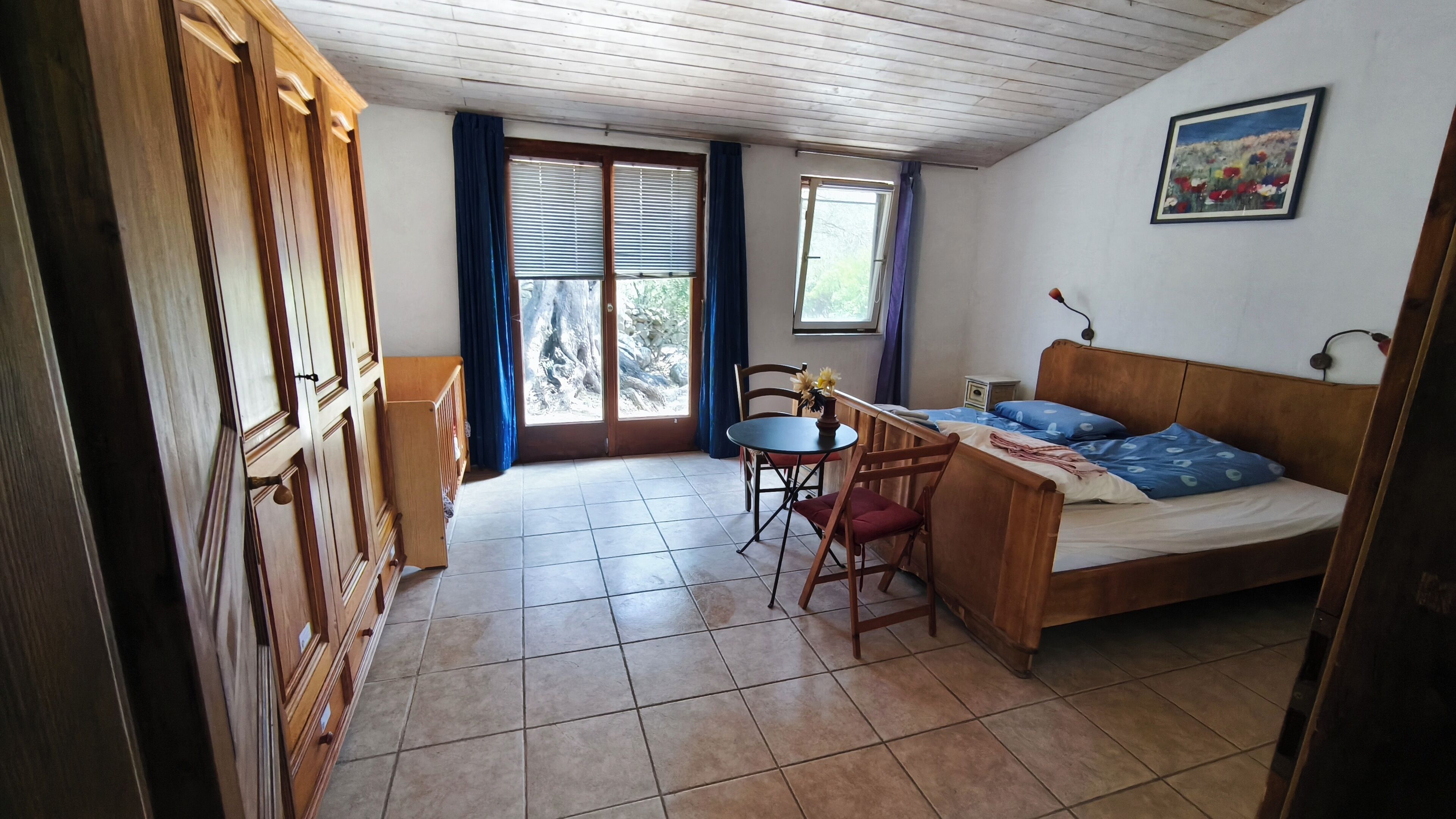 A room with a bed, a small table with chairs, a wooden wardrobe, and a window with curtains.