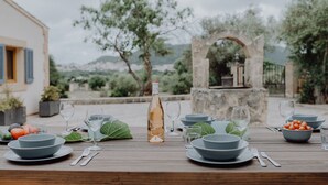 A wooden table set for a meal with plates, bowls, glasses, and a bottle of wine, set against a backdrop of a stone archway and greenery. - Small but nice finca with great views and pool near Cala Ratjada (Capdepera)
