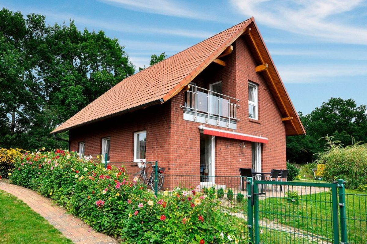 A two-story brick house with a sloped roof, a balcony, and a garden with flowers.