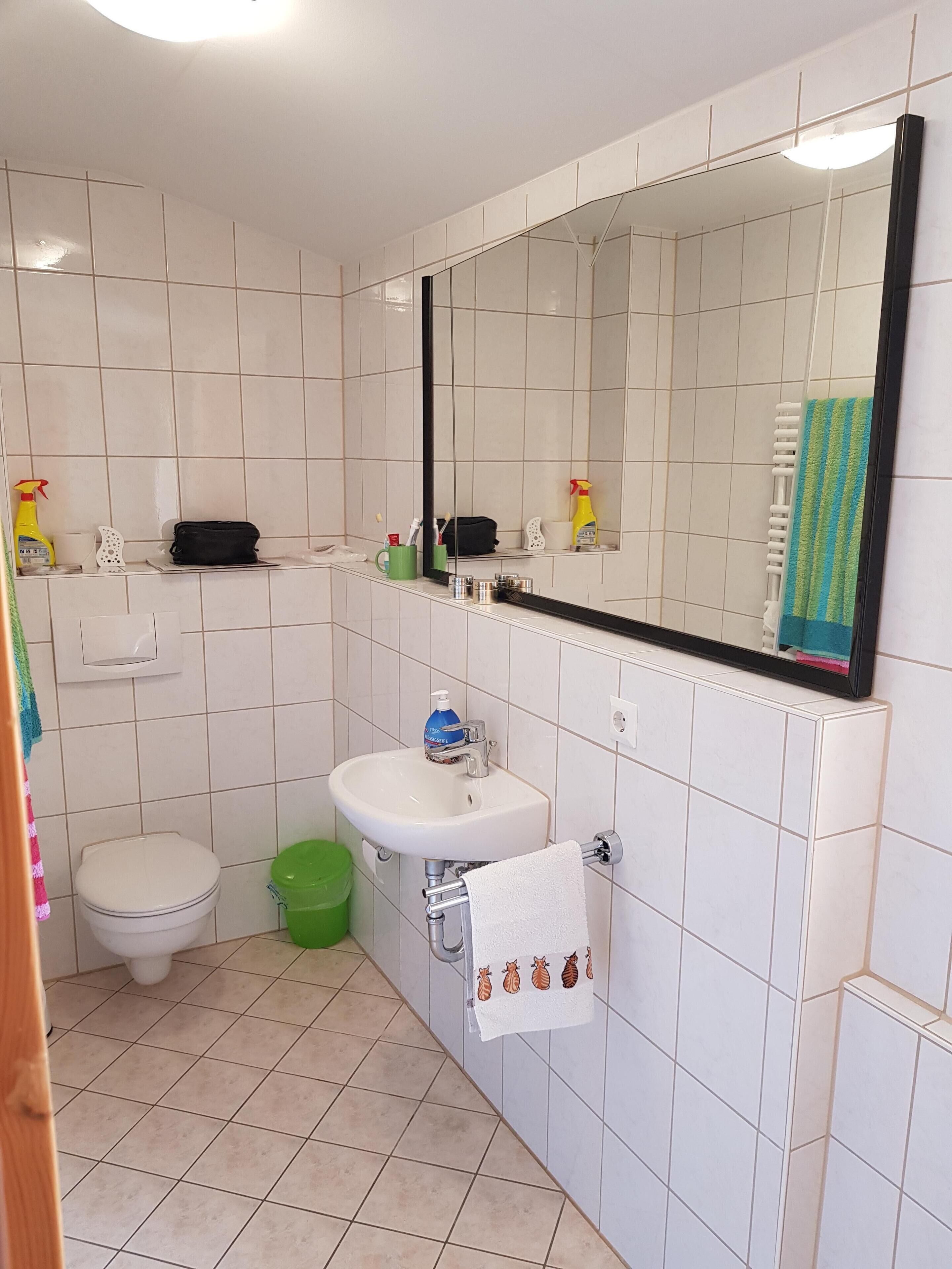 A bathroom with a toilet, sink, and mirror. There is a green bucket and a towel rack with a towel.