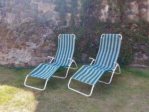 Two striped lounge chairs with metal frames and white armrests, positioned on grass against a stone wall with climbing plants.