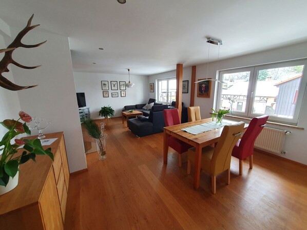 A modern living room with a dining area, wooden flooring, and large windows.