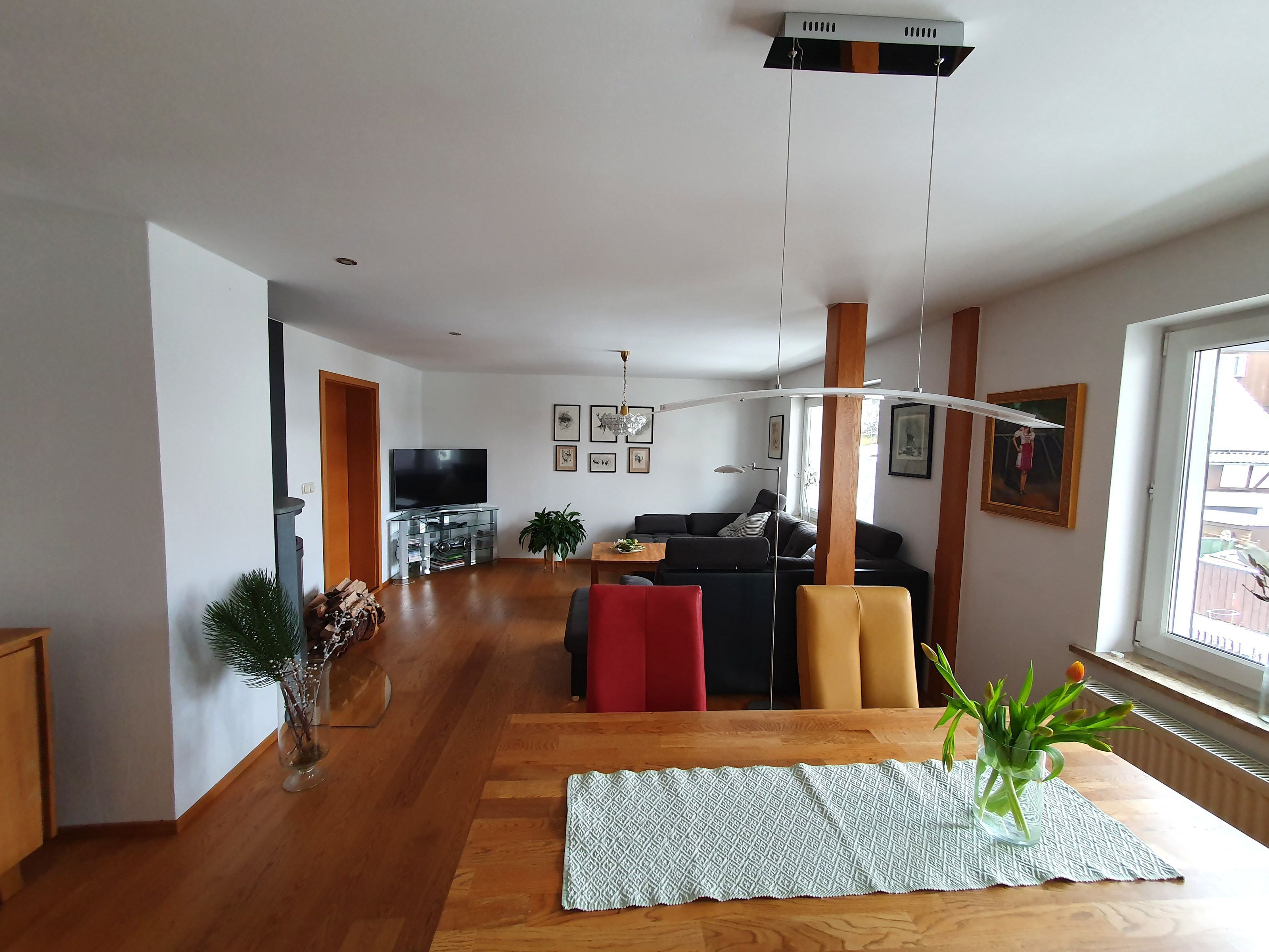 A spacious living area with a wooden floor, a TV on a glass stand, a sofa, a red chair, and a dining table with a placemat.