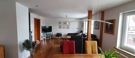 A spacious living area with a wooden floor, a TV on a glass stand, a sofa, a red chair, and a dining table with a placemat.