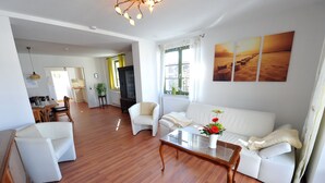 A spacious living room with a white sofa, a coffee table with a vase of flowers, wooden flooring, and a large window with curtains. - Comfortable 4-star roof terrace apartment, quiet and central, 4 adults + 1 baby (Rostock)