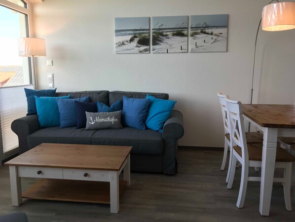 A living room with a grey sofa, blue cushions, a wooden coffee table, a white dining table with chairs, and a beach-themed wall art.