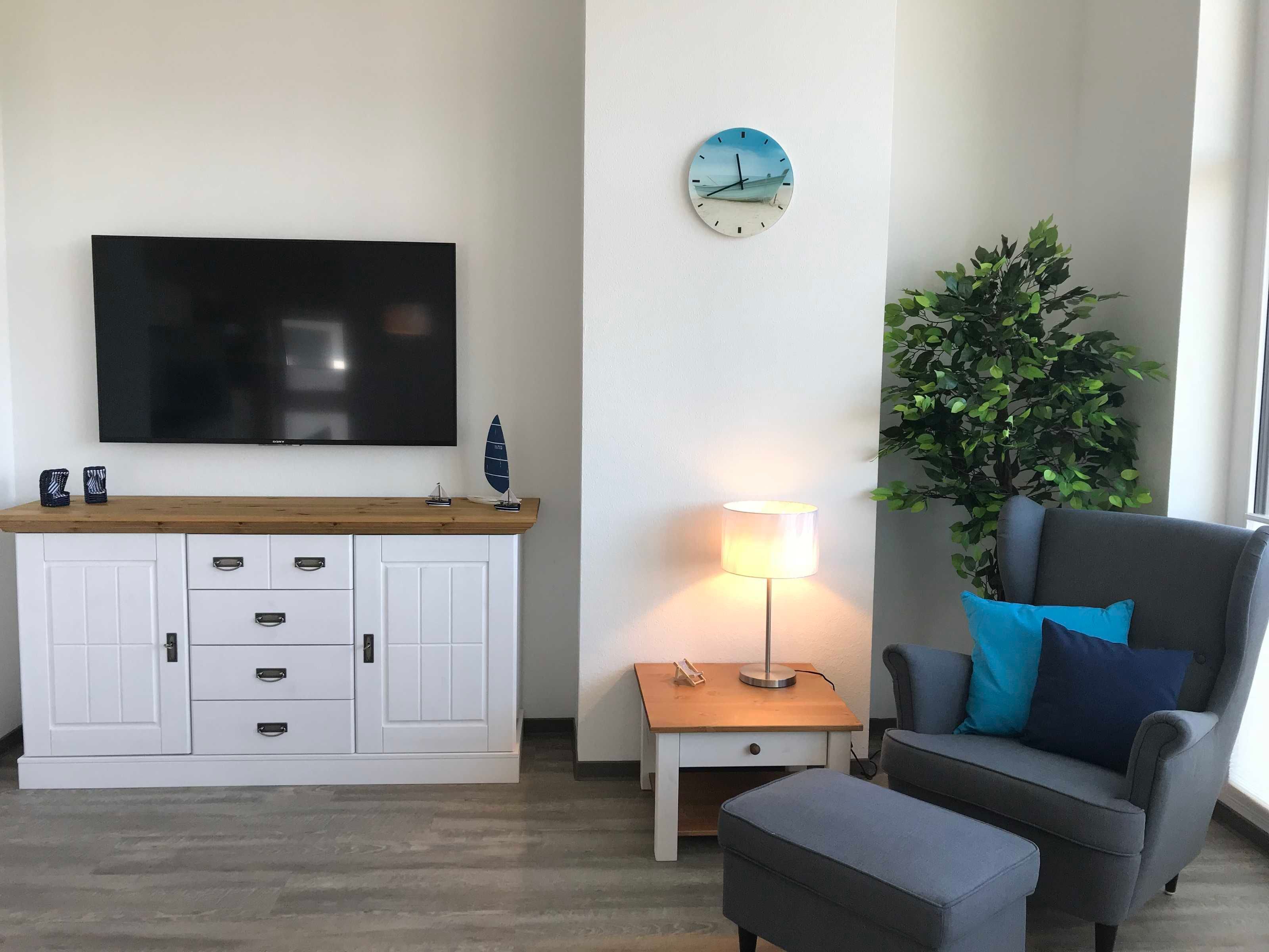 Ein Wohnzimmer mit Flachbildfernseher, Holzschrank, einer Uhr, einer Lampe, einem Blumentopf und einem grauen Sofa mit blauen Kissen.