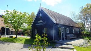 En mørk bygning med skrå tag, et skilt der siger "Fiskershus", samt en lille have med bænk og træ.