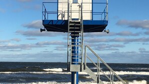 Ein Rettungsschwimmturm mit einer Metalltreppe, die zu einer blau-weißen Struktur führt, mit Blick auf den Ozean.