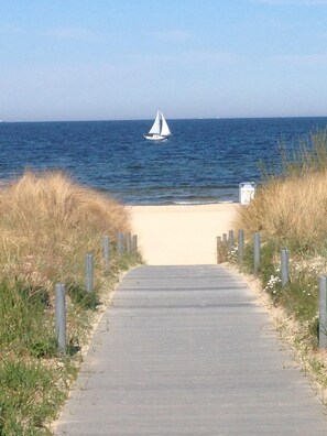 A paved pathway leading to a sandy beach with a sailboat in the distance.