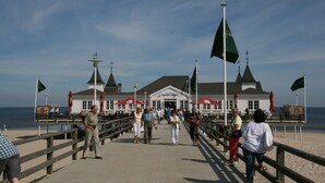 Eine Mole mit einem Gebäude am Ende, mit einer Flagge, einem Fahnenmast und Blick auf den Strand.
