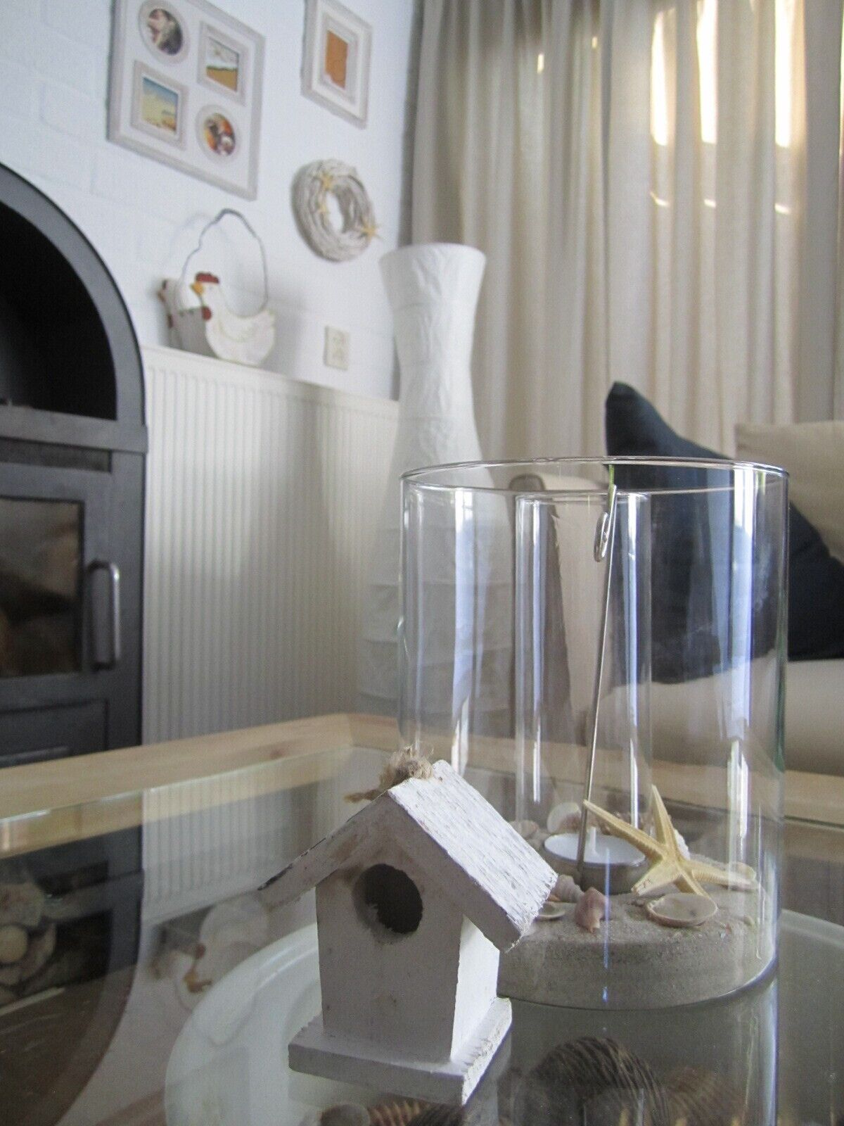 A clear glass birdhouse with a starfish and seashells inside, placed on a glass table.