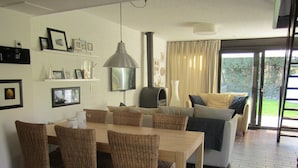A modern living room with a dining area, featuring wicker furniture, a large flat-screen TV, and a view of the outdoors.