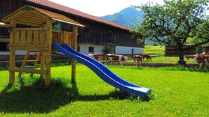 Une cabane en bois avec un toboggan, située dans un champ d’herbe, avec une grange en arrière-plan et des vaches qui paissent.