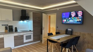 A modern kitchen with white cabinets, a built-in oven, and a coffee maker. There's a dining area with a wooden table and black chairs. A flat-screen TV is mounted on the wall displaying various channels.