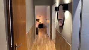 A hallway with wooden flooring, a white door on the left, a white door at the end, and a mirror on the right wall.